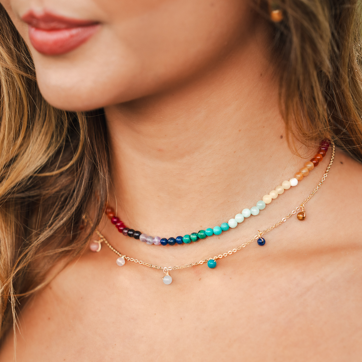 Model wearing a necklace stack. Necklaces include 4mm multicolor stone healing necklace and a 4mm multicolor stone dewdrop charm healing necklace on a gold chain