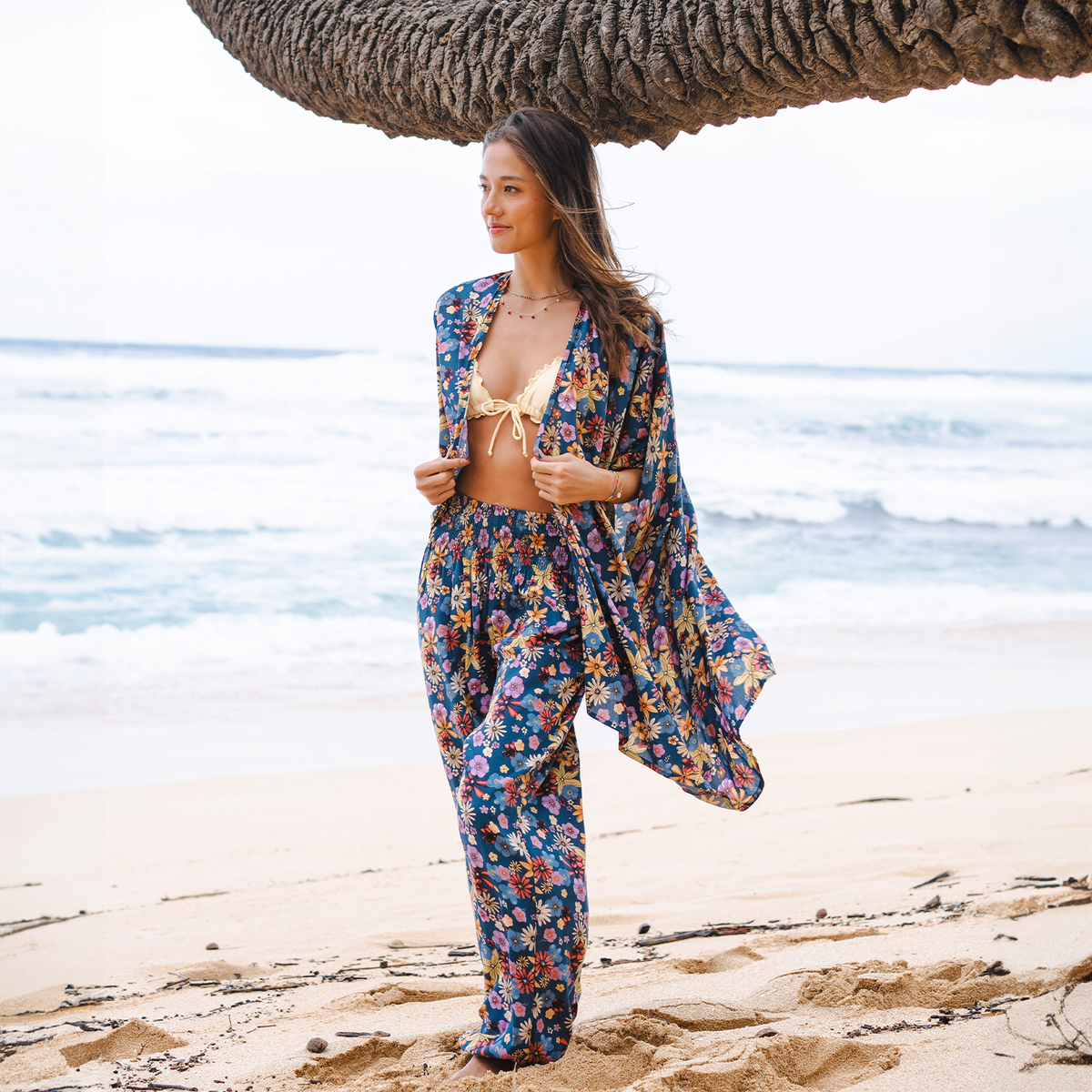 Model standing on the beach wearing blue, pink, orange and yellow floral print harem pants and a matching kimono