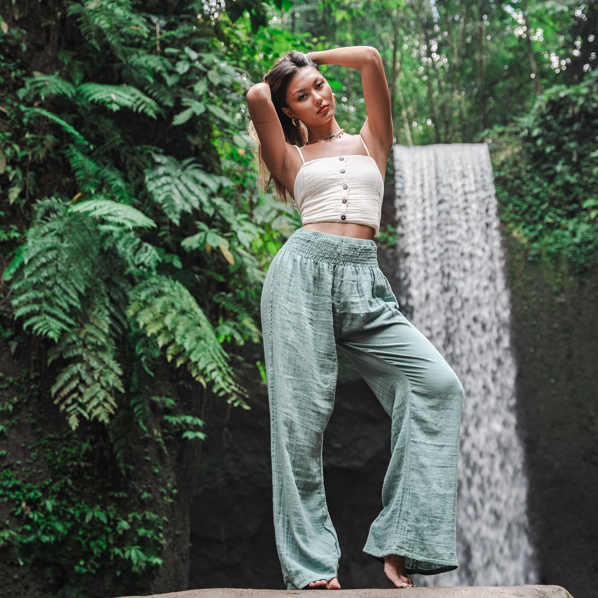 Model standing in front of waterfall wearing a white cotton tank top and sage wide leg cotton pants