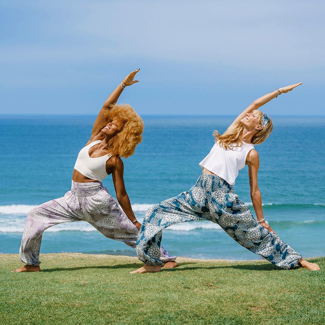 Two women doing yoga poses in front of the ocean. One is wearing deep teal harem pants with a white mandala paisley print and the other is wearing lavendar harem pants with a mandala paisley print