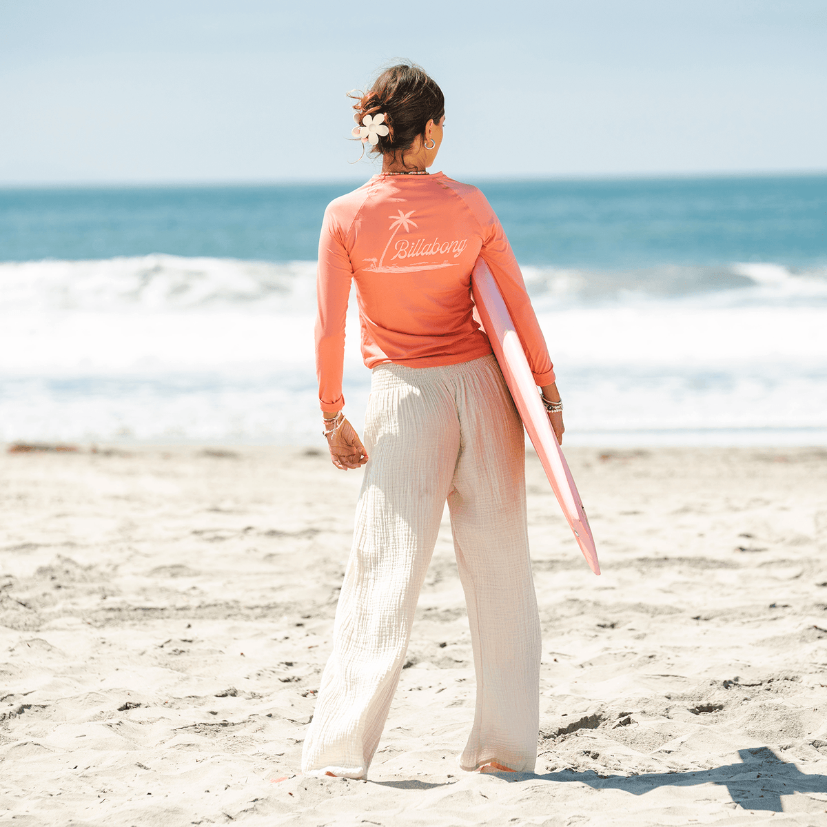 A girl standing on the beach and holding a surfboard while wearing cream wide leg cotton pants