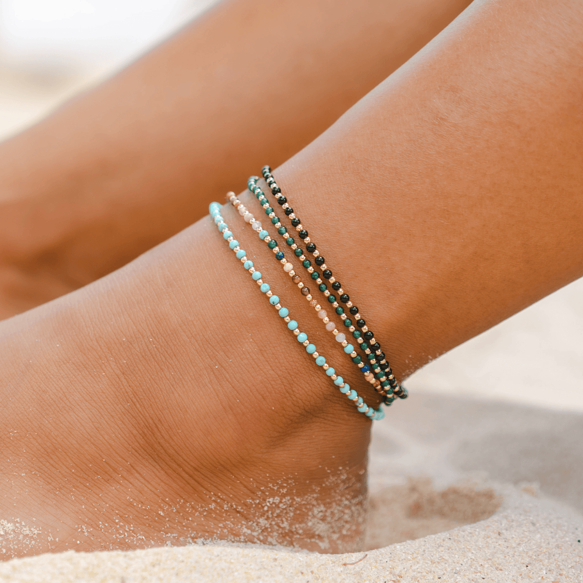 Model wearing a stack of four healing anklets. The anklets include a 2mm turquoise stone and gold bead healing anklet, a 2mm multicolor stone and gold bead healing anklet, a 2mm dark green stones and gold bead healing anklet and a 2mm onyx stone and gold bead healing anklet.