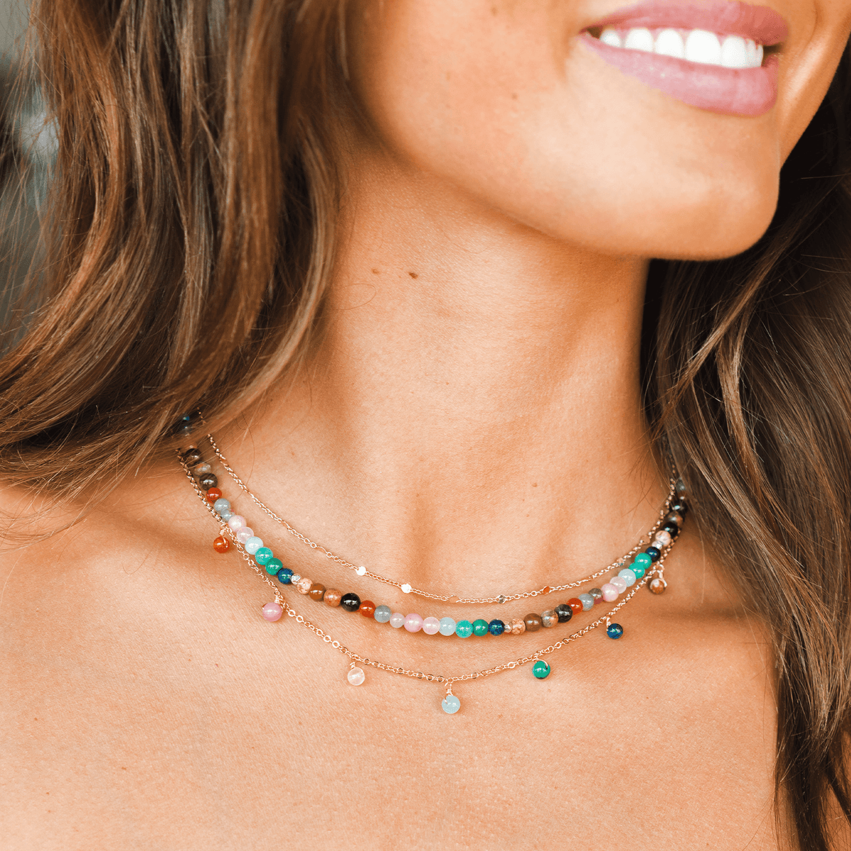 Model wearing a stack of three healing necklaces. The necklaces include a 4mm multicolor stone healing necklace, a gold chain necklace and a gold chain healing necklace with multicolor stone dewdrop charms