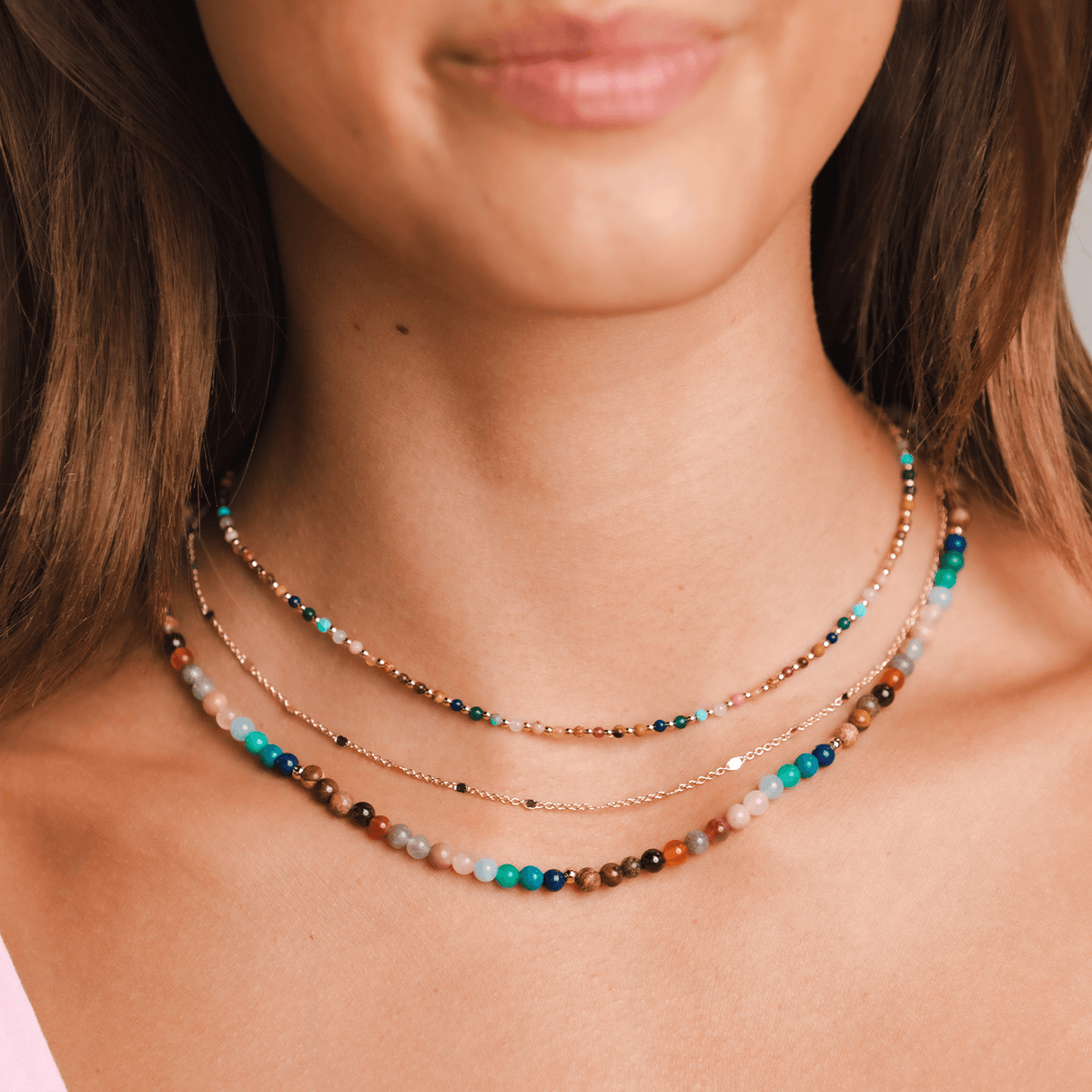 Model is wearing a stack of three necklaces. One is a 4mm multicolor stone necklace, the other is a dainty gold chain necklace and the last one is a dainty 2mm multicolor stone and gold bead necklace