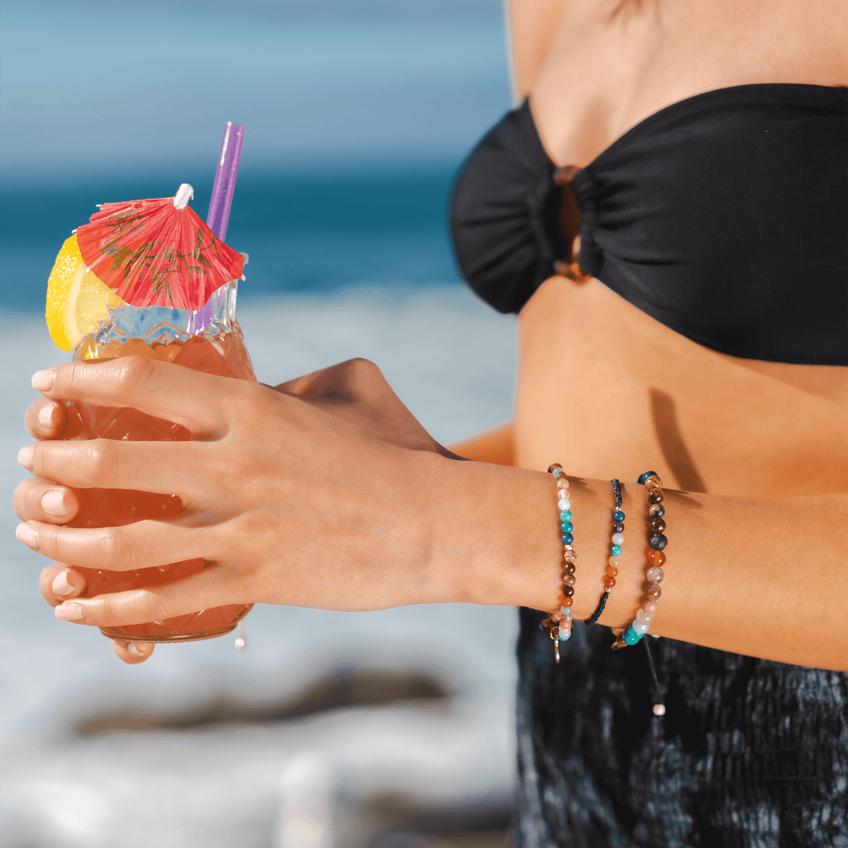 Woman at beach holding drink wearing 4mm Bracelet made up of healing stones including Jade, Jasper, Amazonite, Quartz, Tiger&#39;s Eye, Garnet, and Unakite.