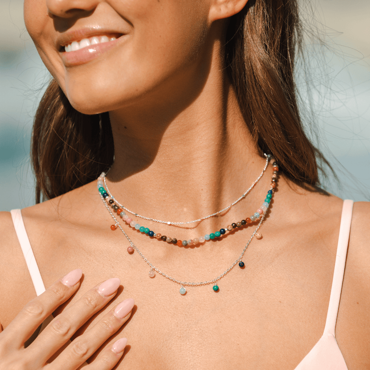 Model wearing a three necklace stack. One is a dewdrop charm style with multicolored beads and a silver chain, the other is a 4mm multicolor bead necklace and the last is a dainty silver chain necklace